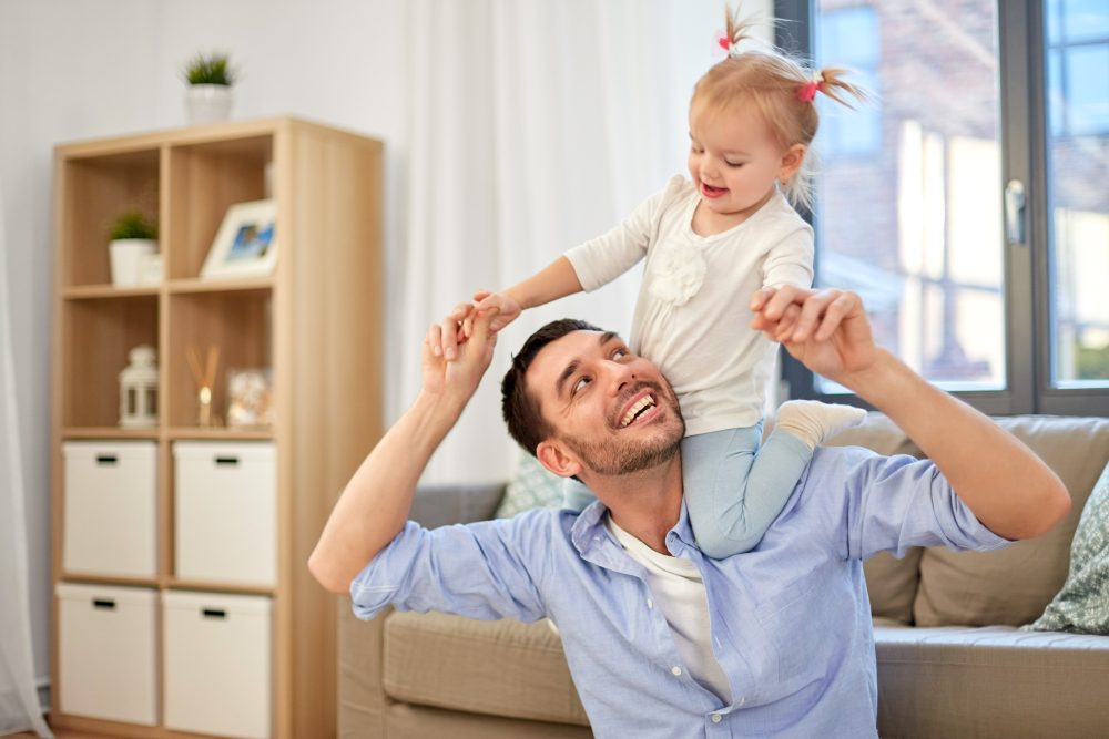 Man in his livingroom with toddler daughter on his shoulders.