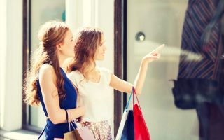 Two women in their late teens or early twenties holding bags on their arms and looking in a store window.