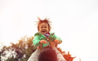 Father is raising happy cheerful toddler son in green jacket up in the air in the park. Toddler boy is playing with father. Happy healthy family, love concept, positive human emotions, feelings.
