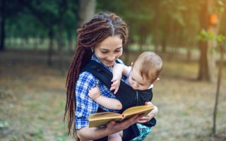 Happy mom with a baby in a sling reading a book in the Park. The concept of children's education in family life