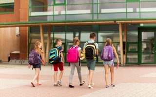 primary education, friendship, childhood and people concept - group of happy elementary school students with backpacks walking outdoors from back