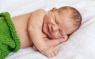 Laughing, smiling little newborn baby boy sleeping in white blanket, lying on bed.