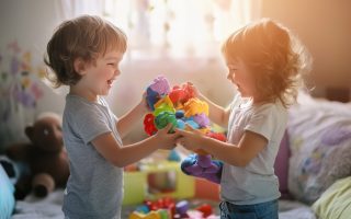 Two young children are happily sharing and playing with vibrant toys in a well-lit bedroom.