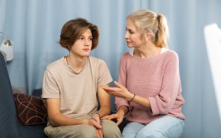 Upset teenager boy after quarrel with his mother in domestic interior, mother consoles son