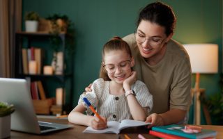 Happy child and adult are sitting at desk. Girl doing homework or online education.