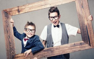 Portrait of two young elegant brothers posing in wooden frame, looking at camera, wearing fashionable eyeglasses and bow tie.