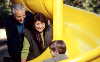 An older adult smiling while holding hands with two young children in a park, with a passerby looking on curiously