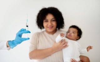 Smiling loving young african american woman holding baby in her arms, looks at doctor hand with syringe on white background, blurred. Mother and child, vaccination, medical health care