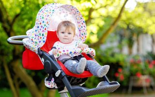 Cute healthy little beautiful baby girl sitting in the pram or stroller and waiting for mom. Happy smiling child with blue eyes. With green tree background. Baby daughter going for a walk with family.