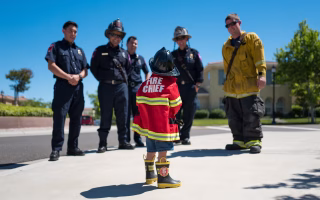 Child in a fire chief costume with real firefighters, symbolizing fire safety lessons.
