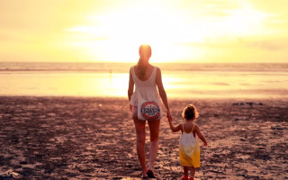 Mother and daughter holding hands while walking on the beach at sunset.