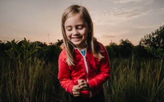 Smiling child in red jacket standing in a grassy field at sunset