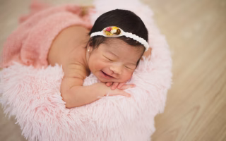 Smiling newborn baby girl with a flower headband lying on a pink fluffy blanket.