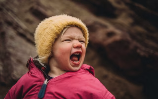 Toddler yelling while wearing a jacket and hat outdoors.