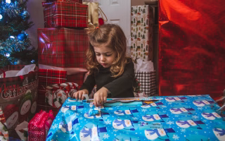 Young girl opening a large Christmas present surrounded by wrapped gifts.