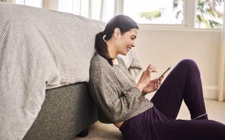 Woman sitting on the floor using her phone, leaning against a bed in a relaxed posture.