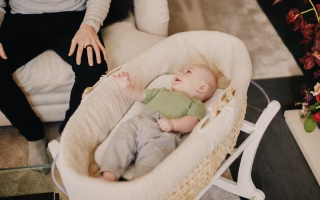 Smiling baby lying in a bassinet next to a seated adult.
