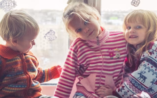 Three young children in colorful sweaters sitting by a window.