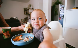 Smiling toddler in a high chair eating banana and finger foods.