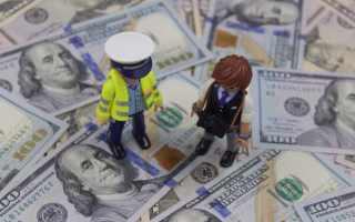 Toy figures of a police officer and photographer standing on a pile of hundred-dollar bills.