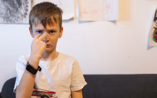 Young boy pointing seriously at his face while standing indoors.
