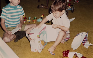 Two young boys sit on a carpeted floor excitedly unwrapping a toy gift box.