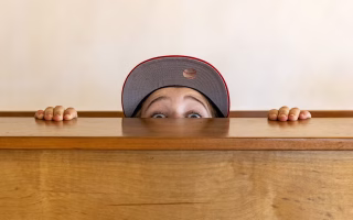 Child in a baseball cap peeking over a wooden surface with wide eyes.