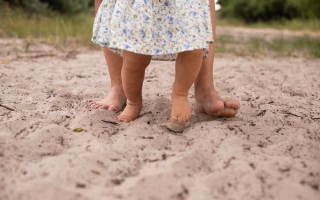 Barefoot toddler walking on tiptoes in sand with adult support.