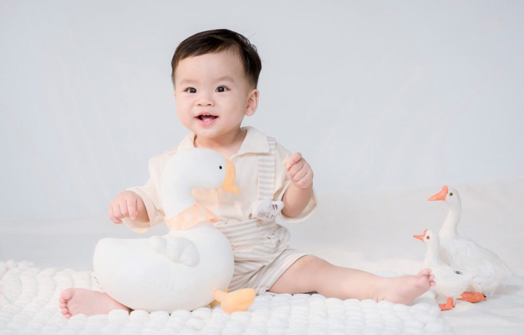 Smiling baby in a white outfit sitting beside toy ducks